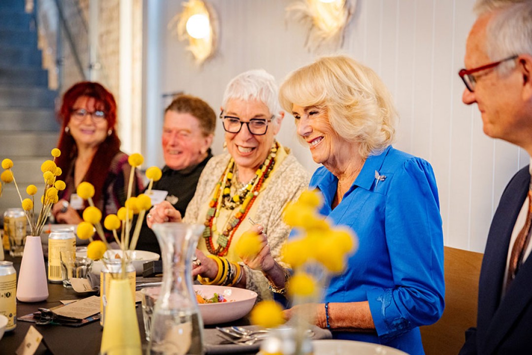 Her Majesty sits, smiling, at a table, with a plate of food in front of her. Other diners, also smiling, sit on either side.