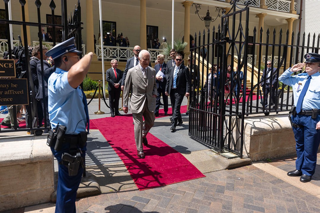 His Majesty walking out of the NSW Parliament on a red carpet being saluted by guards and followed by security.