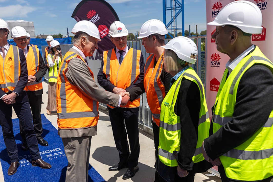 His Majesty wears a high-vis vest and a hard hat among a group of people wearing the same. He is shaking hands with one of them. 