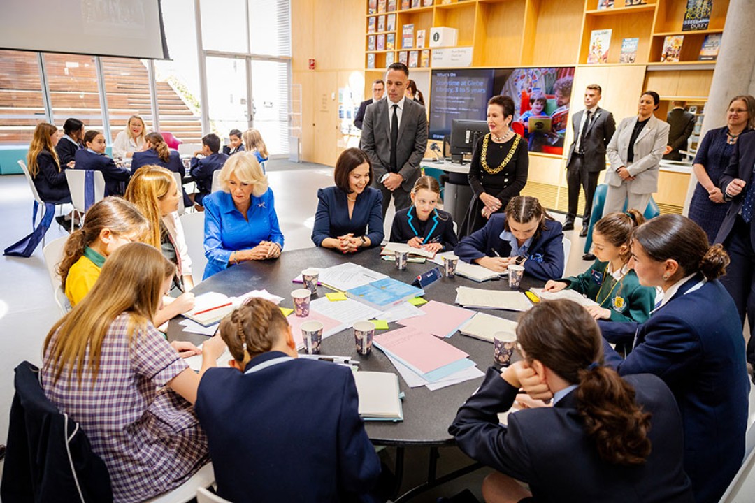 Her Majesty sits at a table with a group of children in school uniform. 