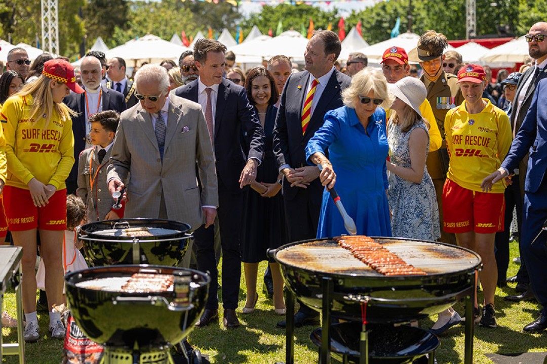 Their Majesties tend snags on barbecues while a crowd looks on. Some of the crowd are wearing Surf Life Saving Australia uniforms.