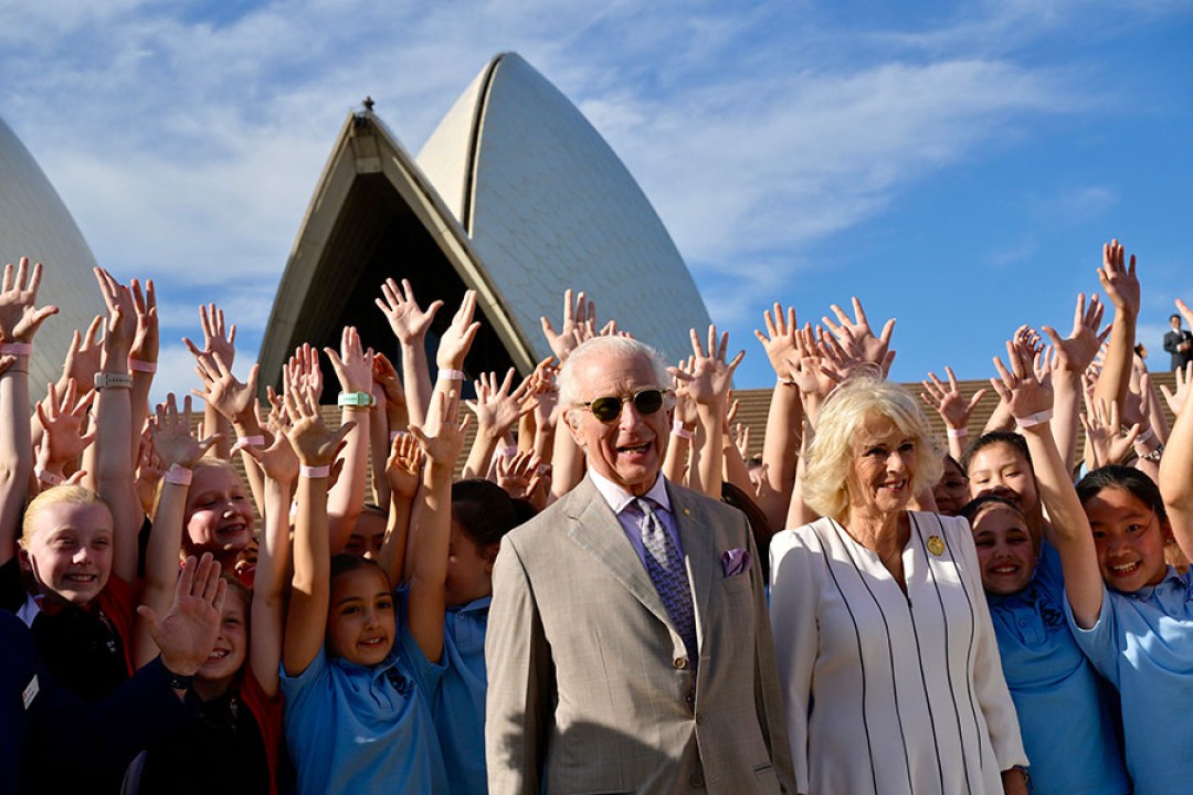 Their Majesties stand with a crowd of children in front of the Sydney Opera House. The children have their arms stretched towards the sky.