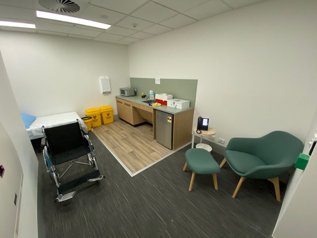 Shows a room with a wheelchair in the foreground, a bed at the back left of the room, a chair to the right of the entry, and bench with a sink, mini fridge, microwave and first aid boxes at the far right of the room.