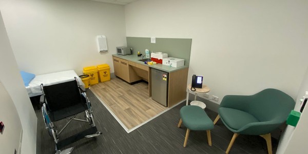 Shows a room with a wheelchair in the foreground, a bed at the back left of the room, a chair to the right of the entry, and bench with a sink, mini fridge, microwave and first aid boxes at the far right of the room.