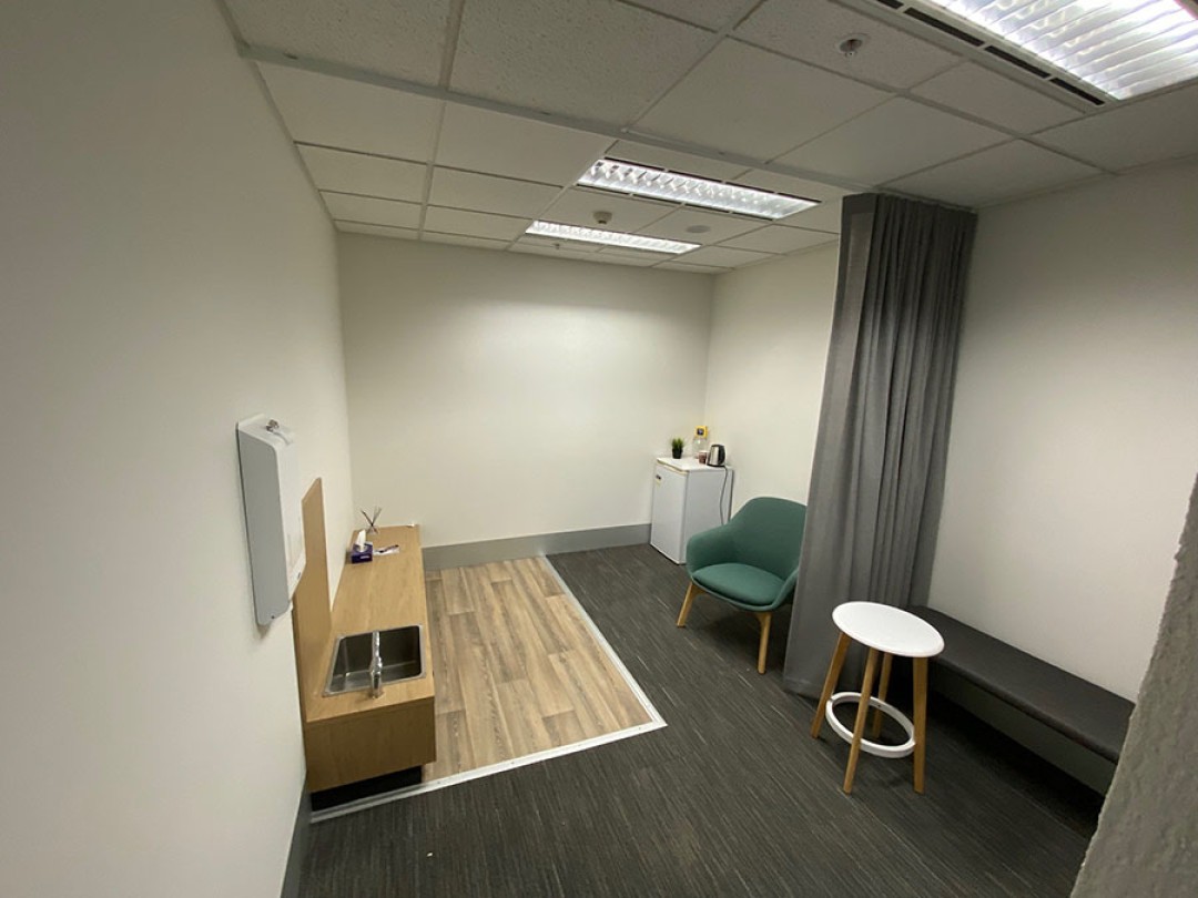 Shows a room with a foot wash sink to the left of the room, a mini-fridge and chair at the back right of the room, and a privacy curtain.