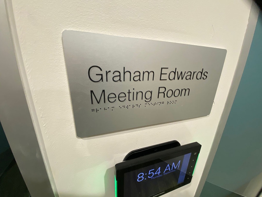 Shows a sign outside a meeting room reading “Graham Edwards Meeting Room,” with braille at the bottom of the sign.