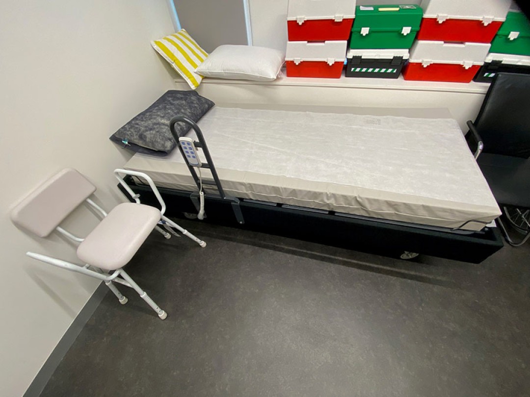 Shows a room with an electric, height adjustable bed at the far end of the room, a chair to the right of the bed, and a collection of first aid boxes.