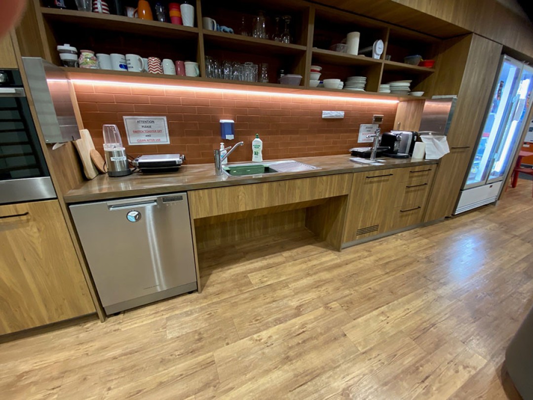 Shows a kitchen area with a refrigerator on the far right wall, with a long kitchen bench that has a wheelchair accessible sink, water station, and kitchen appliances.