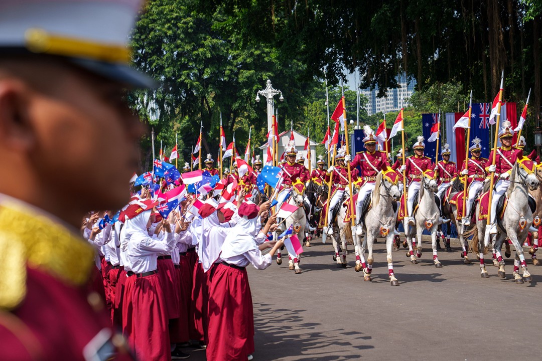 Welcome ceremony in Jakarta. A crowd of people holding flags watch a procession of people on horse back.