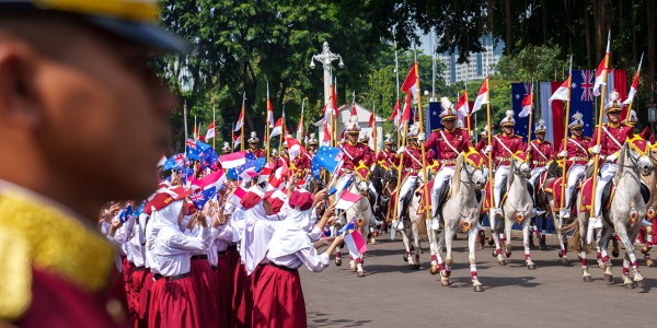 Welcome ceremony in Jakarta. A crowd of people holding flags watch a procession of people on horse back.