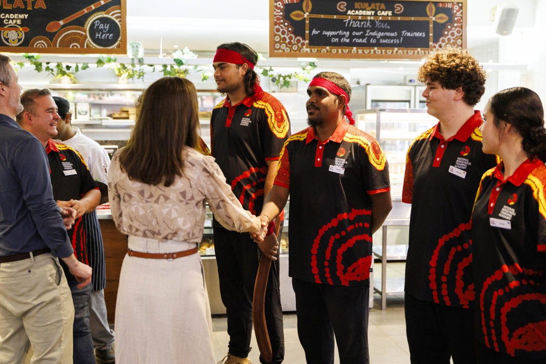 A group of people stand inside the "KULATA ACADEMY CAFE". Text above reads "Thank You, for supporting our Indigenous Trainees on the road to success!". Her Majesty The Queen of Denmark shakes hands with an Indigenous man wearing "NATIONAL INDIGENOUS TRAINING ACADEMY" attire.