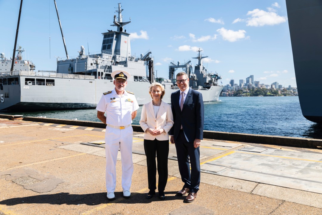 President von der Leyen in a light jacket stands on a dock with two men, including a naval officer in uniform, with large naval ships and a city skyline in the background.