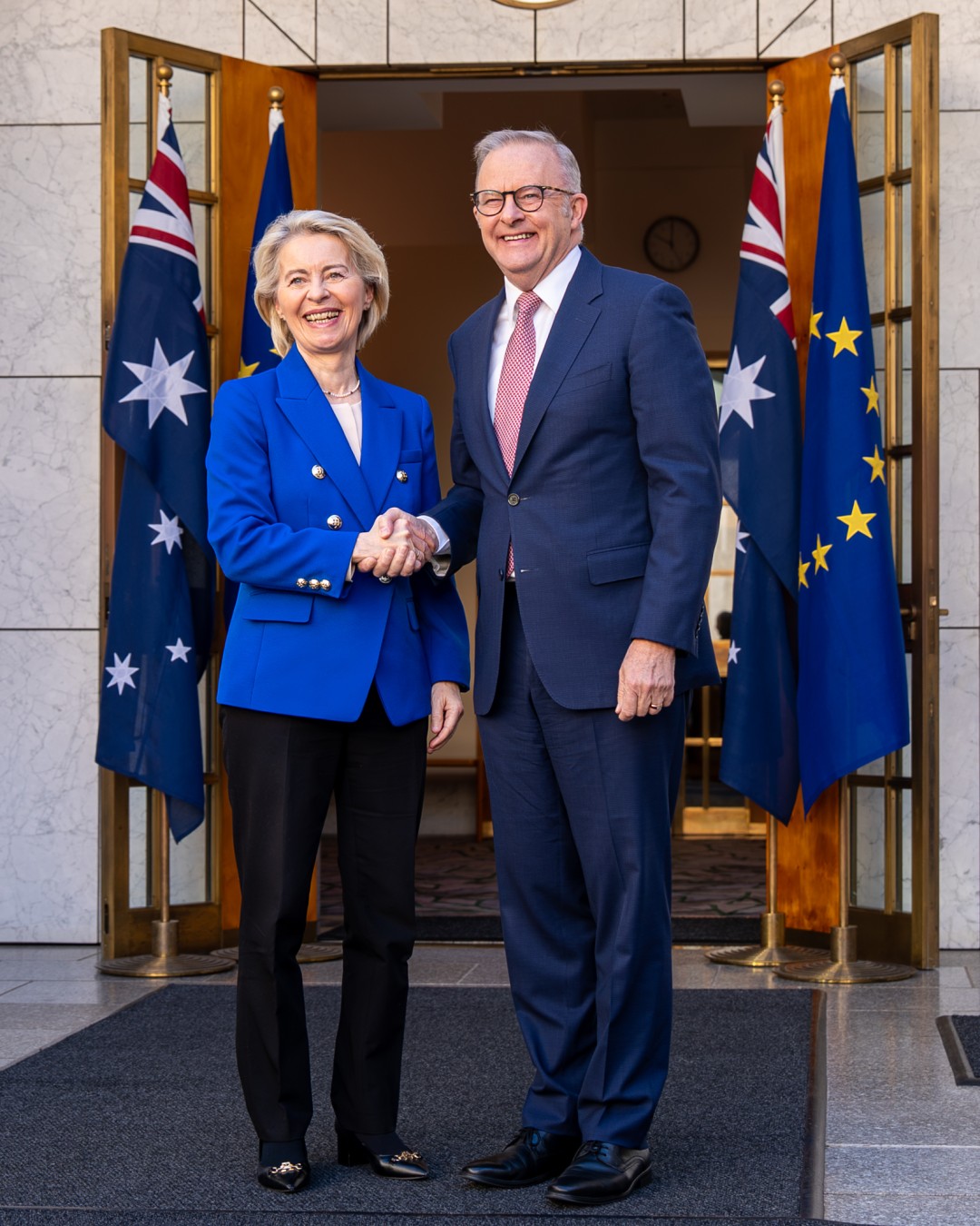 President von der Leyen and Prime Minister Albanese shake hands in front of Australian and European Union flags at a building entrance.