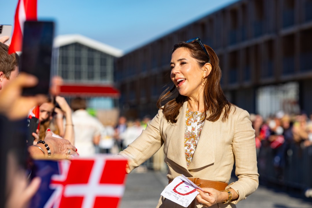 Her Majesty The Queen of Denmark stands outdoors, smiling and reaching out to a crowd holding Danish flags and phones. She holds a child's drawing. A modern building is in the background.