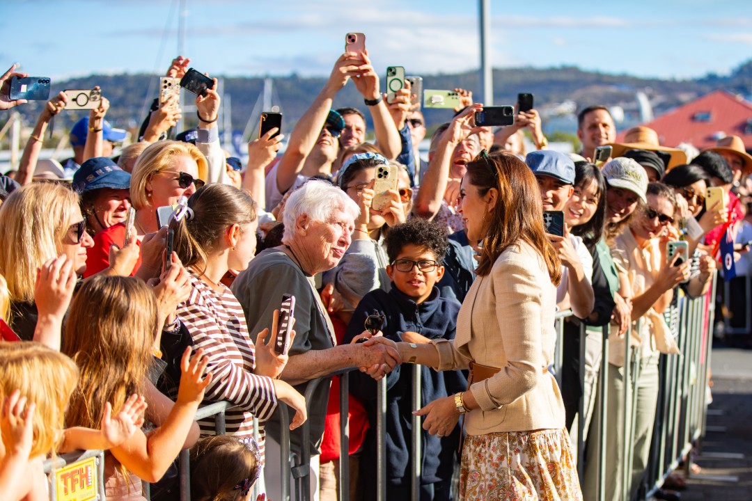 Her Majesty The Queen of Denmark shakes hands with an older woman as crowds behind a barrier photograph the event. Her Majesty wears a blazer and patterned skirt. The crowd holds up phones to capture the moment.