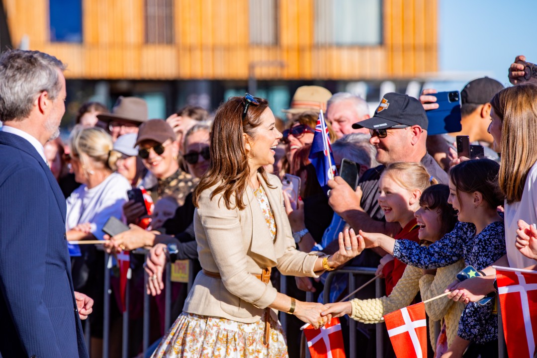 Her Majesty The Queen of Denmark, with auburn hair, smiles, reaching to shake hands with children holding Danish flags, behind a barrier. People behind them use phones to capture the moment asHis Majesty The King of Denmark looks on.