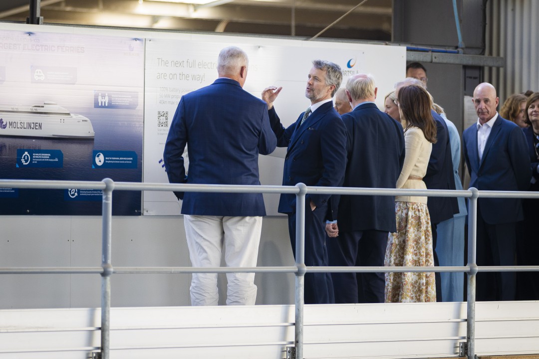 A group with His Majesty The King of Denmark stands by a poster that reads "The next full electric ferries are on the way! 100% battery powered, 50 MW propulsion, 40kn max speed". Text on another poster reads "LARGEST ELECTRIC FERRIES MOLSLINJEN". They are at INCAT Tasmania.