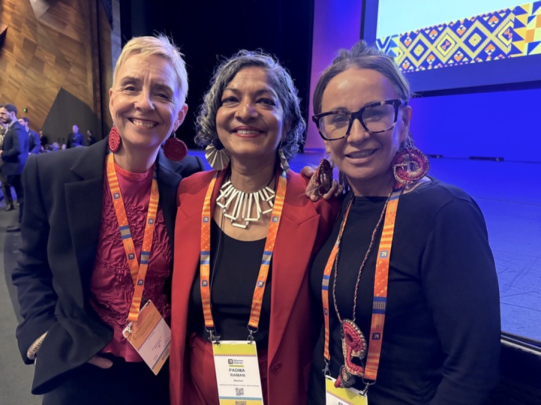 Micaela Cronin, Padma Raman PSM and Sue-Ann Hunter wearing bright orange lanyards, standing in front of a blue lit backdrop.