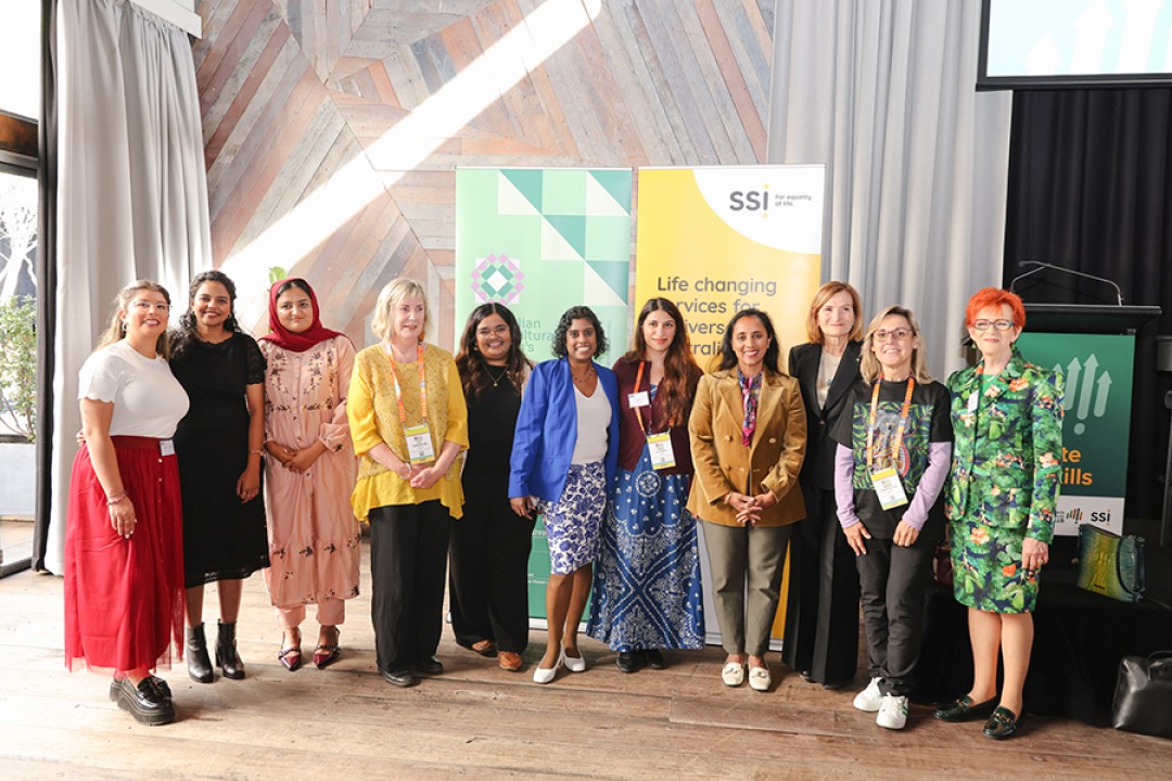 Photograph of ten people standing together in front of banners for Settlement Services International and Australian Multicultural Women's Alliance.