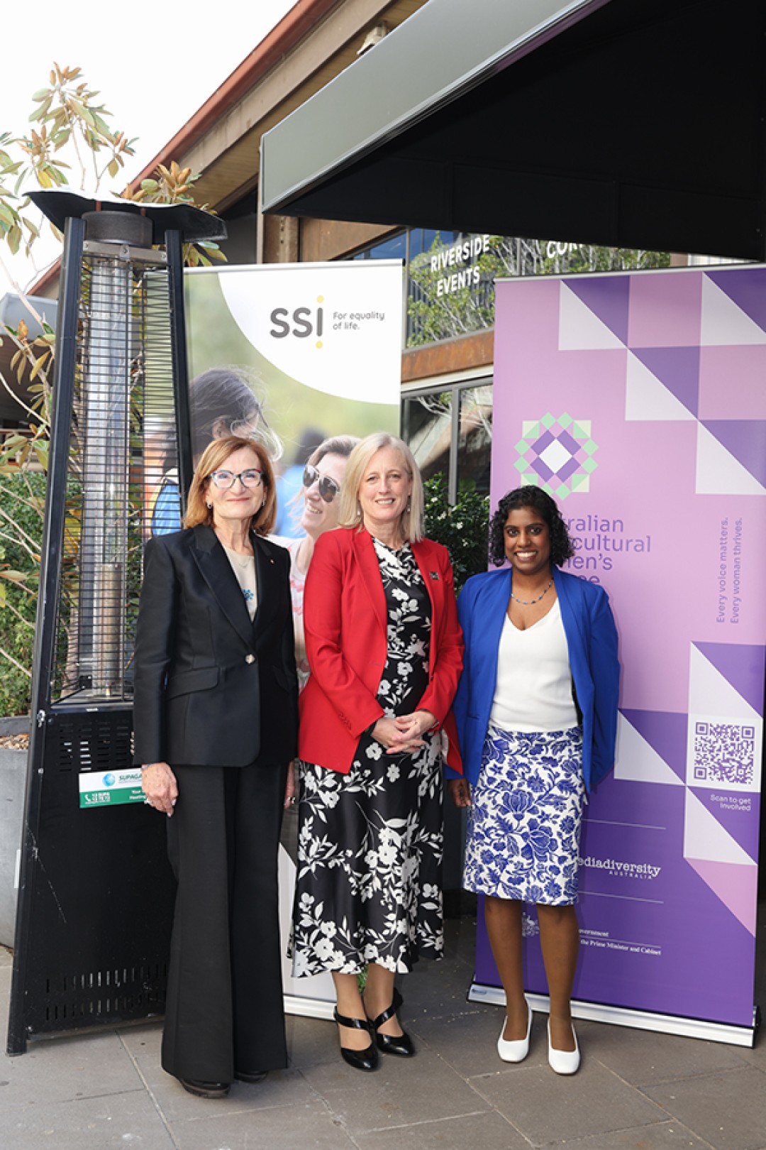 Violet Roumeliotis AM, Senator the Hon Katy Gallagher and Malini Raj, standing in front of two vertical banners promoting Settlement Services International and the Australian Multicultural Women's Alliance.