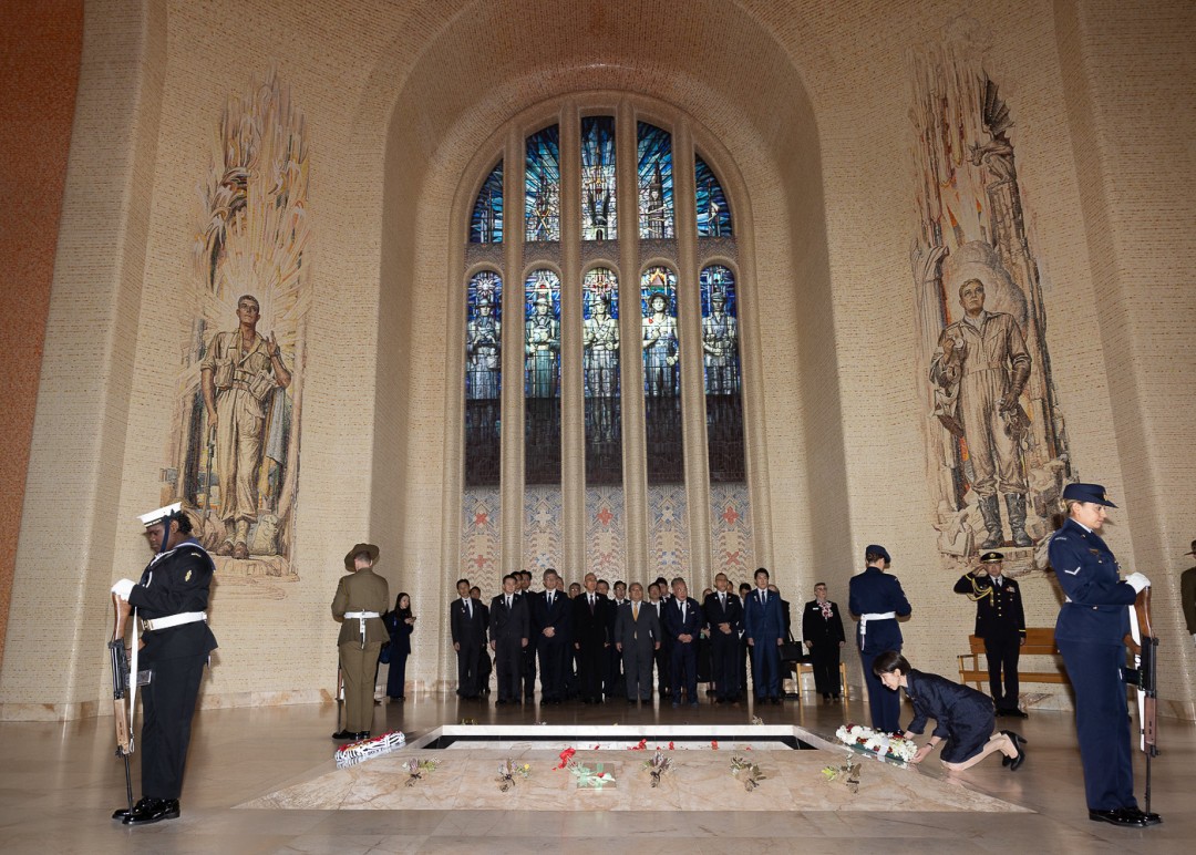 A formal ceremony takes place inside the Australian War Memorial with military personnel and attendees gathered beneath a large memorial window.