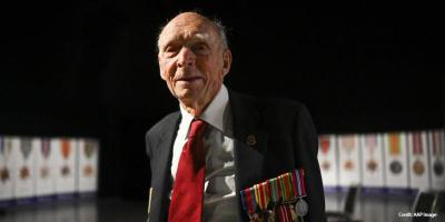 An elderly man in suit and red tie and medals on his chest looks to camera. In the background is a display of medals.