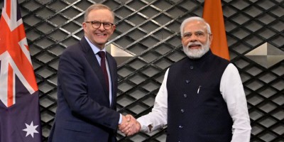 Prime Minister Albanese dressed in a dark suit and tie shakes hands with Prime Minister Narendra Mohdi dressed in Indian formal wear. They stand in front of two flags, two seats and a small table. In the background is a wall painted like steel mesh.