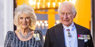 King Charles III in formal wear including medals on a black suit stands next to The Queen Consort in a formal black and white long-sleeved gown with necklace, earrings and a crown.