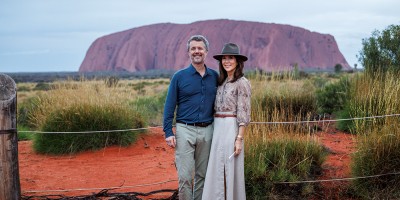 Their Majesties the King and Queen of Denmark stand side by side, smiling, in front of Uluru in Australia. The red sandstone monolith rises in the background under an overcast sky, while dry grasses and red earth surround the couple in the foreground. The King wears a navy shirt and light trousers, and the Queen wears a long skirt, patterned blouse, and a wide-brimmed hat.