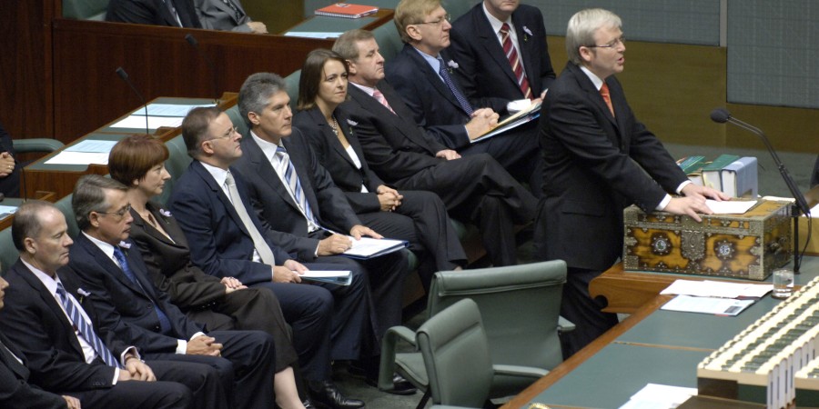 Former Prime Minister Kevin Rudd stands at the dispatch box in the House of Representatives Chamber to deliver the National Apology to Australia’s Indigenous Peoples. He wears a black suit, red tie and glasses. Behind him are other members of parliament seated on green benches, including Deputy Prime Minister Julia Gillard and Foreign Minister Stephen Smith. They are dressed in business attire.