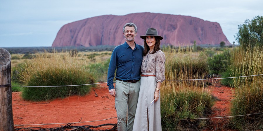 Their Majesties the King and Queen of Denmark stand side by side, smiling, in front of Uluru in Australia. The red sandstone monolith rises in the background under an overcast sky, while dry grasses and red earth surround the couple in the foreground. The King wears a navy shirt and light trousers, and the Queen wears a long skirt, patterned blouse, and a wide-brimmed hat.